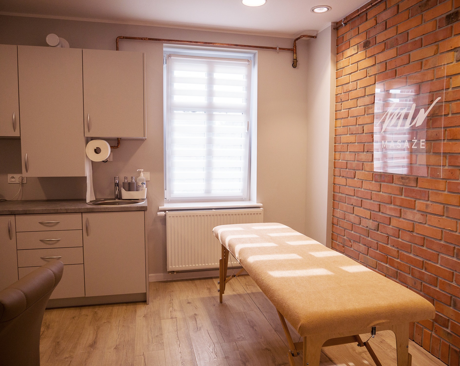 Jasny gabinet z łóżkiem do masażu, ceglaną ścianą z logo, kuchennym aneksem i oknem z żaluzjami.
