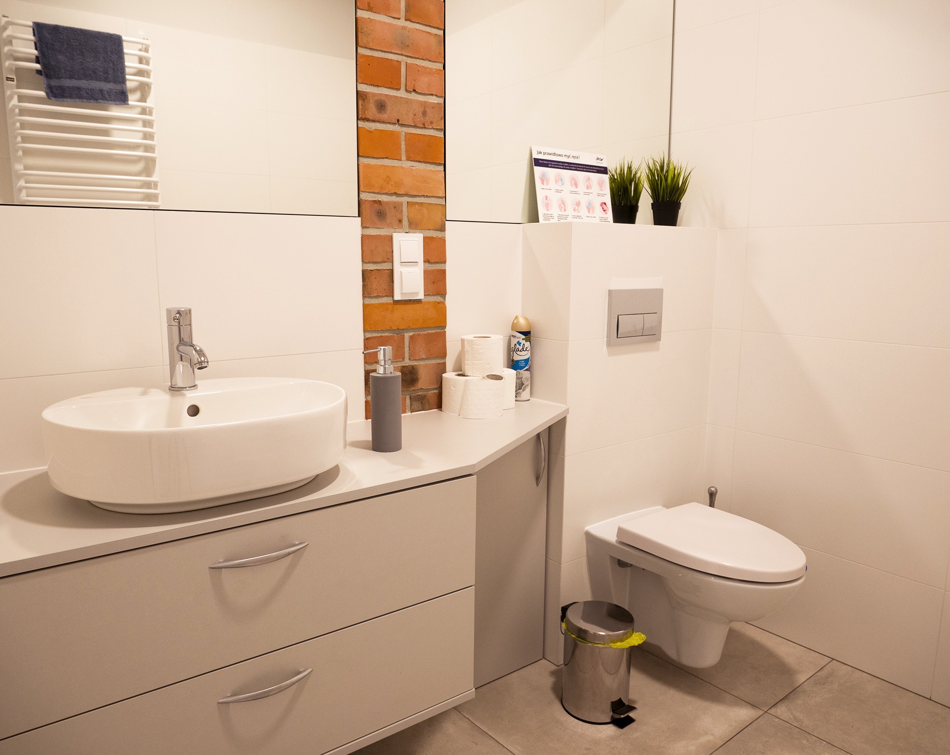 Nowoczesna łazienka z umywalką nablatową, szarą szafką z szufladami, toaletą podwieszaną, koszem na śmieci ze żółtą folią, fragmentem ściany z cegły, plakatem z instrukcją mycia rąk oraz ręcznikiem...