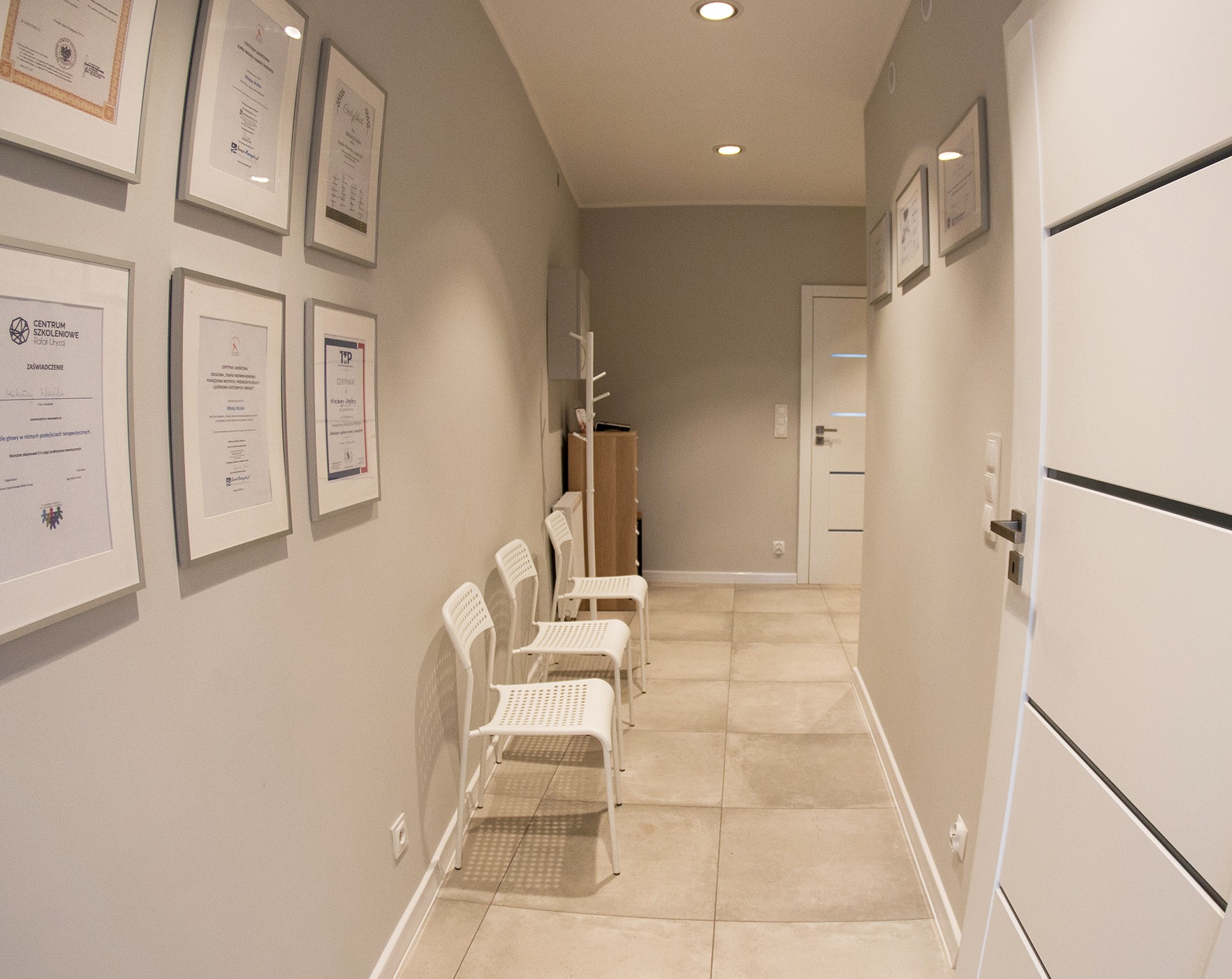 Korytarz w gabinecie z czterema białymi krzesłami dla pacjentów, ściany ozdobione certyfikatami i dyplomami w ramkach, jasna podłoga i oświetlenie punktowe.