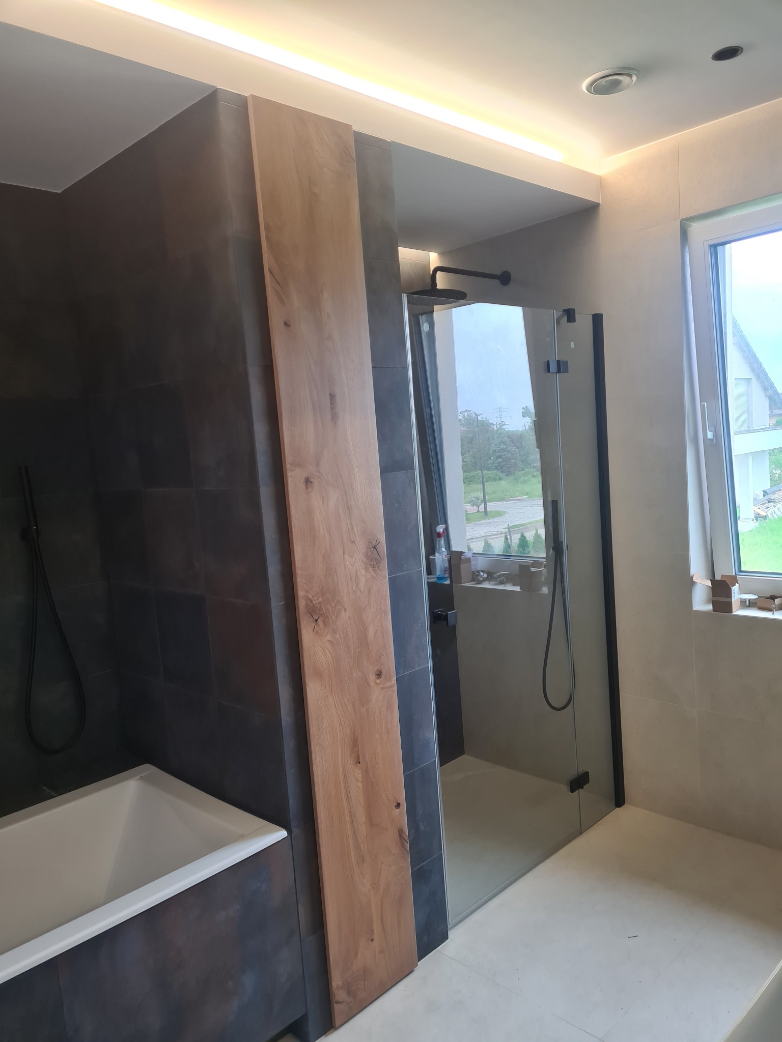 Nowoczesna łazienka z kabiną prysznicową typu walk-in, wykończona ciemnymi płytkami i elementami z drewna, z widoczną wanną i oknem z widokiem na dom.