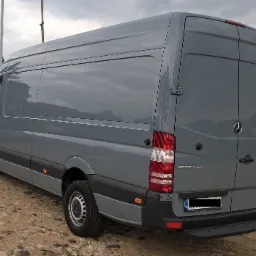Szary samochód dostawczy Mercedes-Benz Sprinter z widocznymi drzwiami tylnymi, oponami i tablicą rejestracyjną, sfotografowany na żwirowym podłożu w pochmurny dzień.
