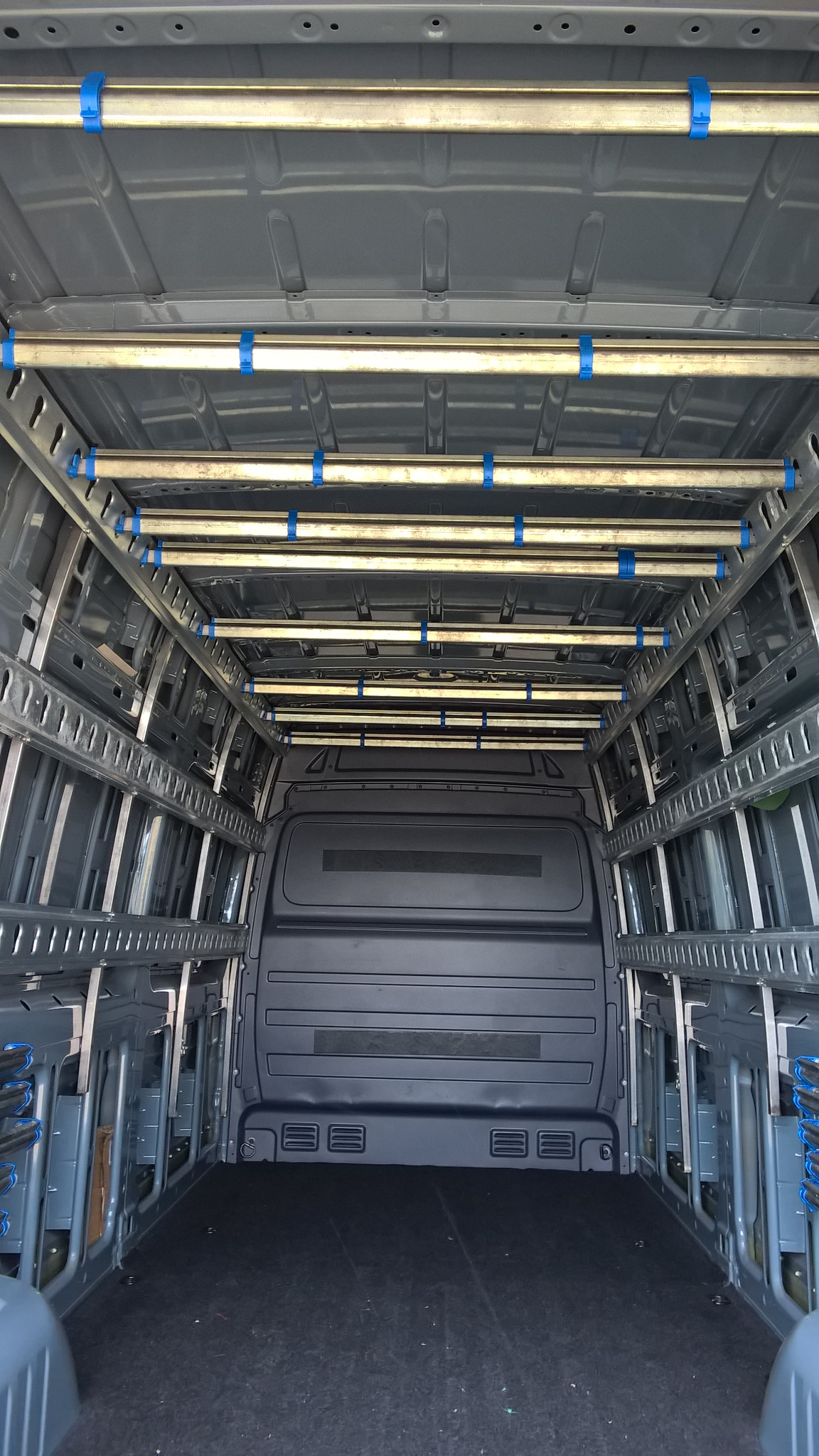 Pusta przestrzeń ładunkowa furgonetki z zamontowanymi metalowymi ramami i poprzeczkami zabezpieczonymi niebieskimi uchwytami, przygotowana do przewozu towarów.