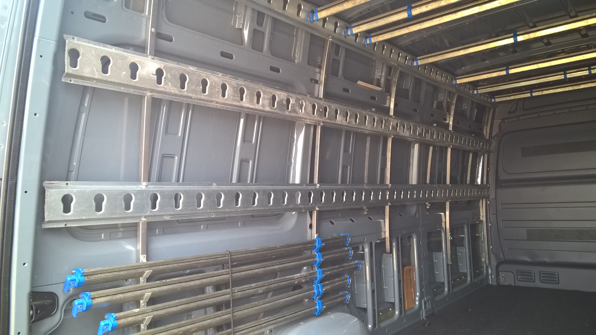 Wnętrze furgonetki z zamontowanymi metalowymi szynami mocującymi i rurami zabezpieczonymi niebieskimi zaciskami, przygotowane do transportu.