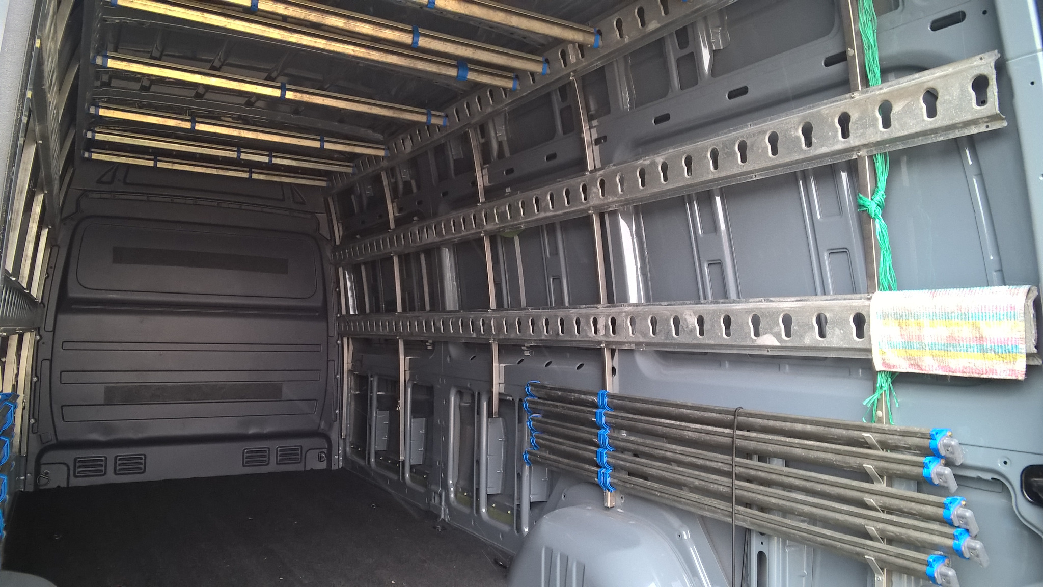 Wnętrze szarego furgonetki z zamontowanymi metalowymi szynami i uchwytami do mocowania ładunku, zabezpieczonymi niebieskimi taśmami, z zielonym sznurkiem i szmatką przywiązana do szyny.