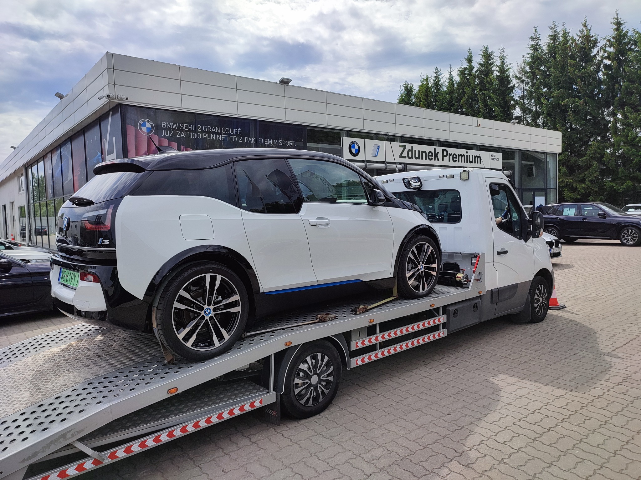 Białe BMW i3 na lawecie przed salonem samochodowym Zdunek Premium, widoczne tablice rejestracyjne i elementy zabezpieczające auto.