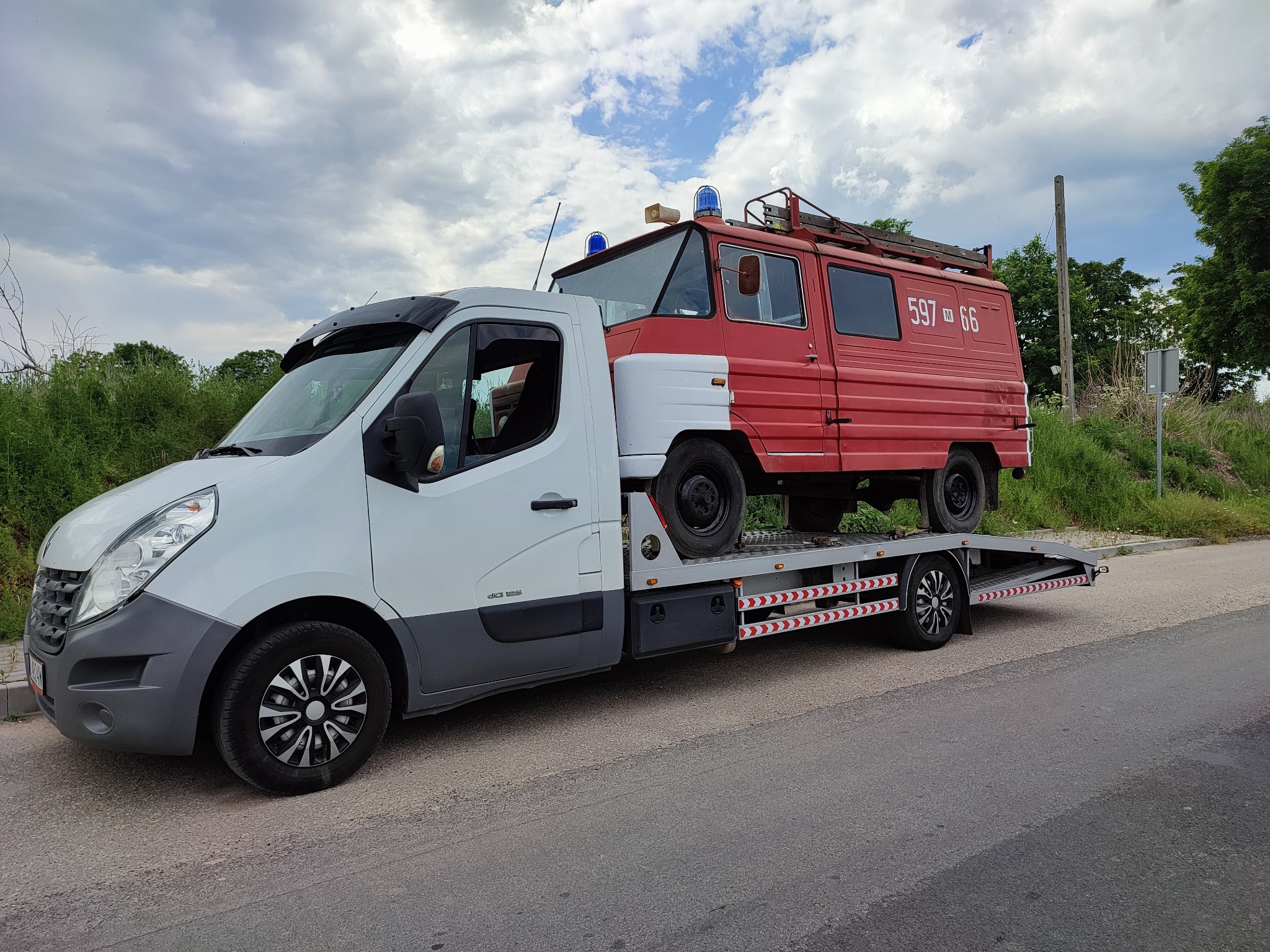 Biała laweta przewozi czerwono-biały wóz strażacki z numerem 597-66 na tle pochmurnego nieba i zieleni.