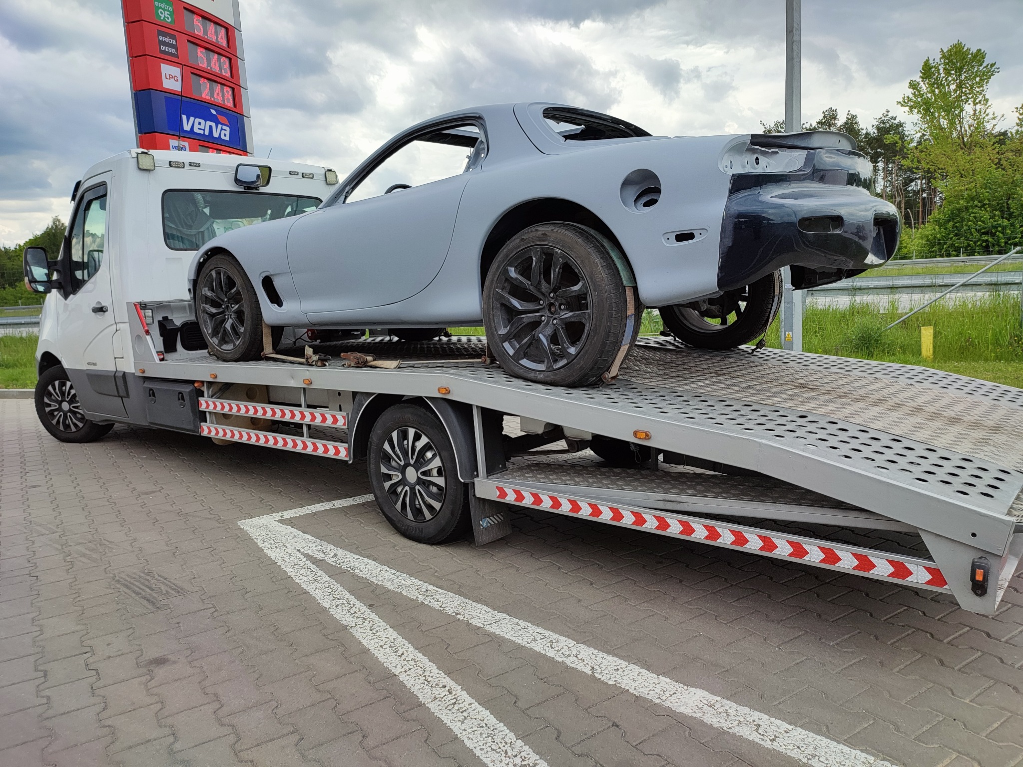 Szara, niekompletna karoseria sportowego samochodu Mazda RX-7 na lawecie przed stacją benzynową.