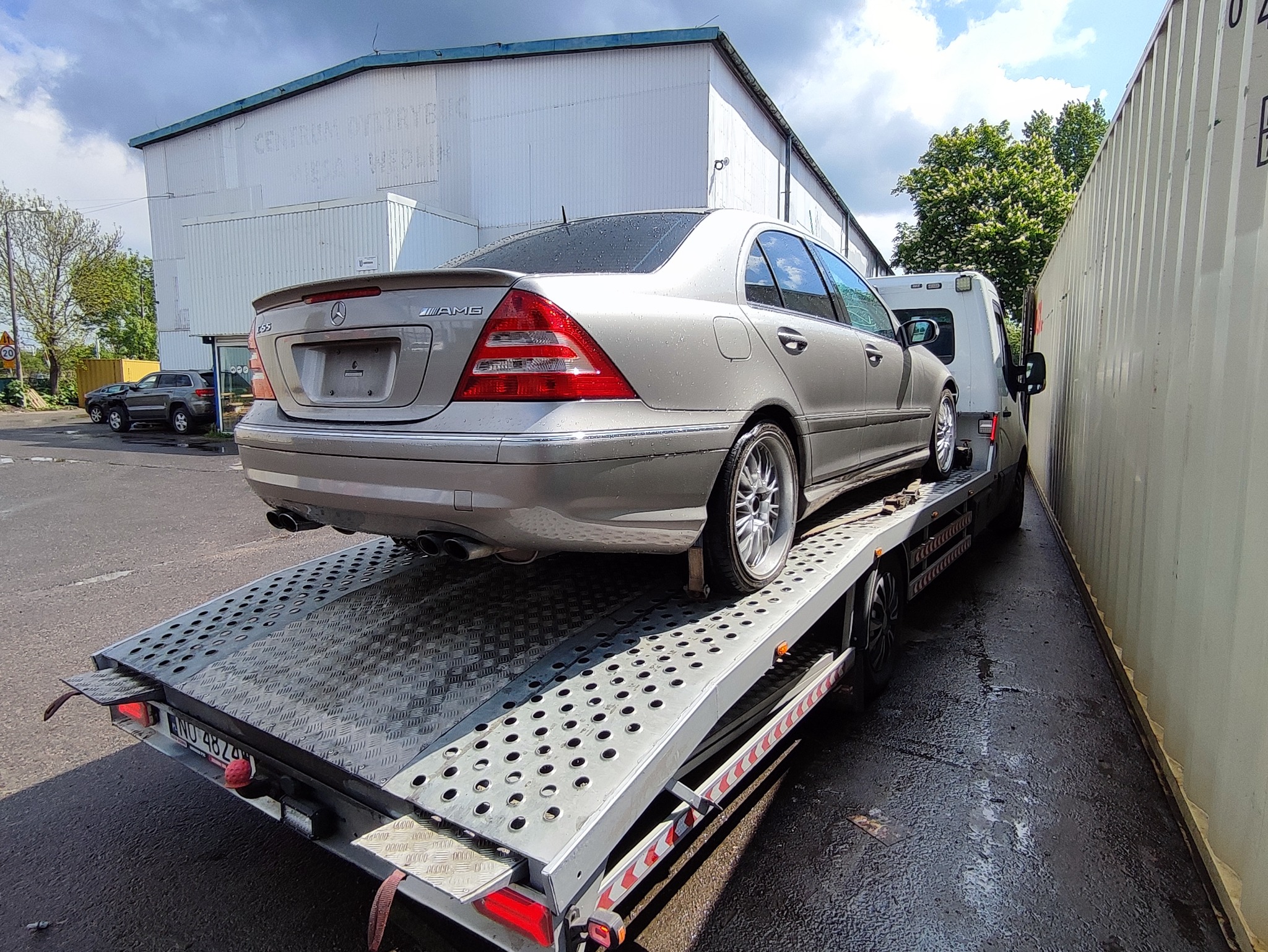 Srebrny Mercedes-Benz C55 AMG na platformie lawety z widocznym fragmentem numeru rejestracyjnego, na tle budynku Centrum Dystrybucji i kontenera.