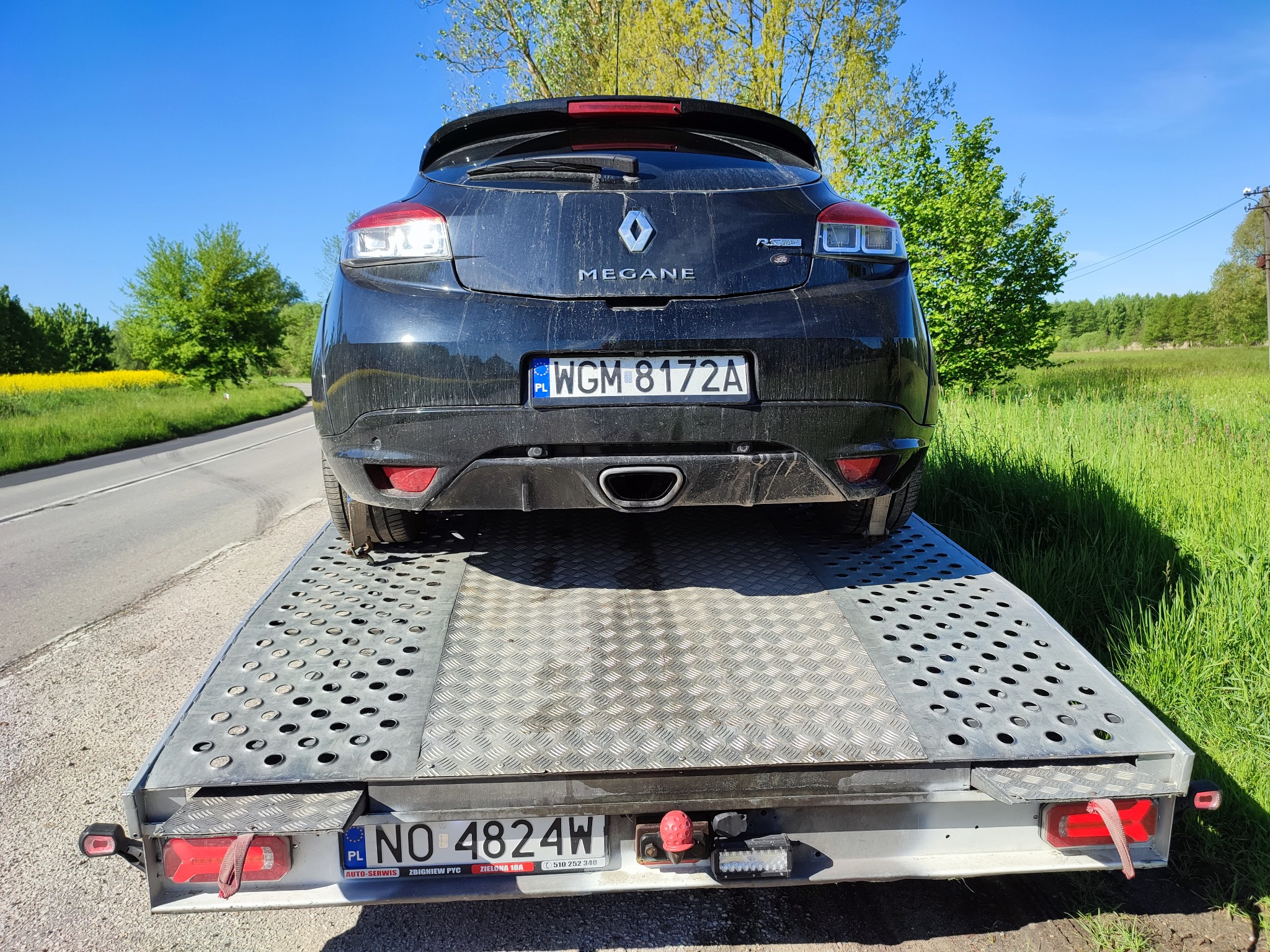 Czarny samochód Renault Megane RS na ażurowej platformie lawety, widok z tyłu, tablice rejestracyjne widoczne na samochodzie i lawecie, słoneczny dzień.