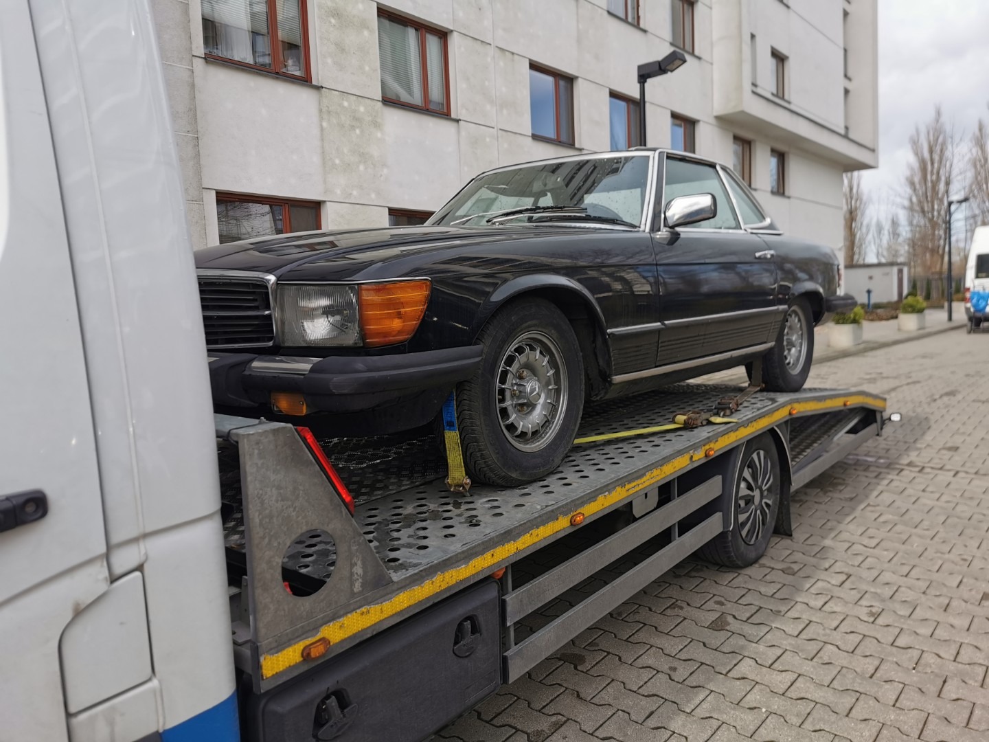 Czarny, klasyczny Mercedes SL na platformie lawety, zabezpieczony pasami transportowymi, na tle bloku mieszkalnego.