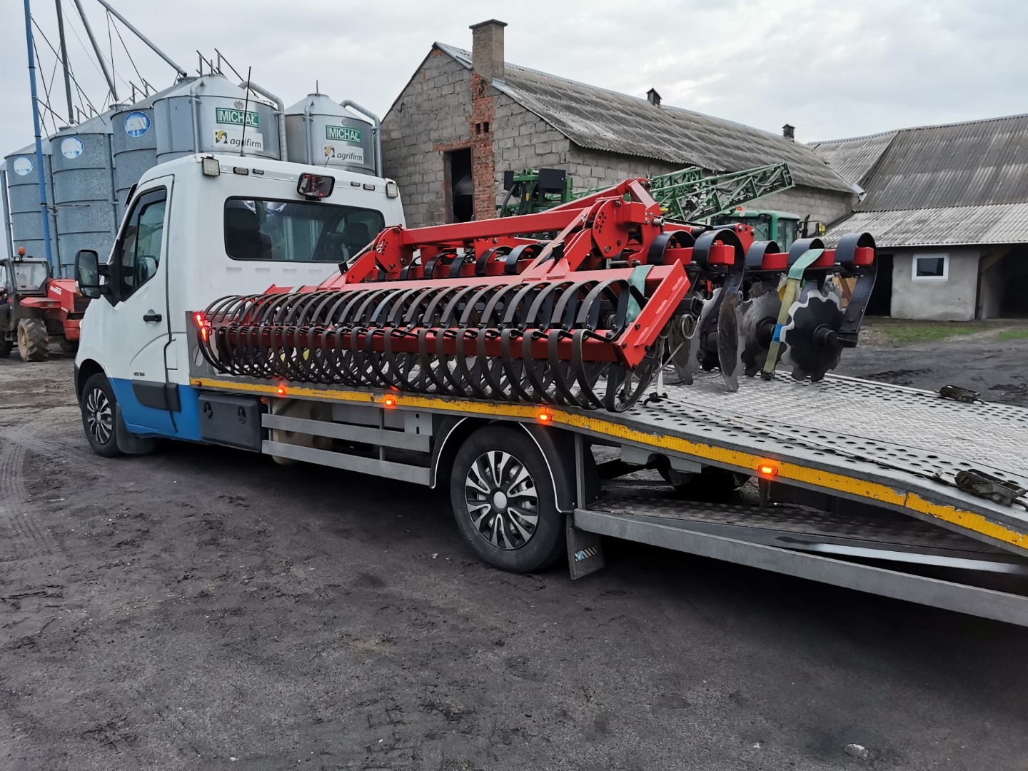 Laweta przewozi czerwony agregat uprawowy na tle zabudowań gospodarczych i silosów.
