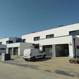 Nowoczesne szeregowe domy jednorodzinne z płaskimi dachami, białą elewacją i szarymi akcentami, częściowo z otwartymi garażami, biały samochód dostawczy na podjeździe, palety budowlane i kostka...