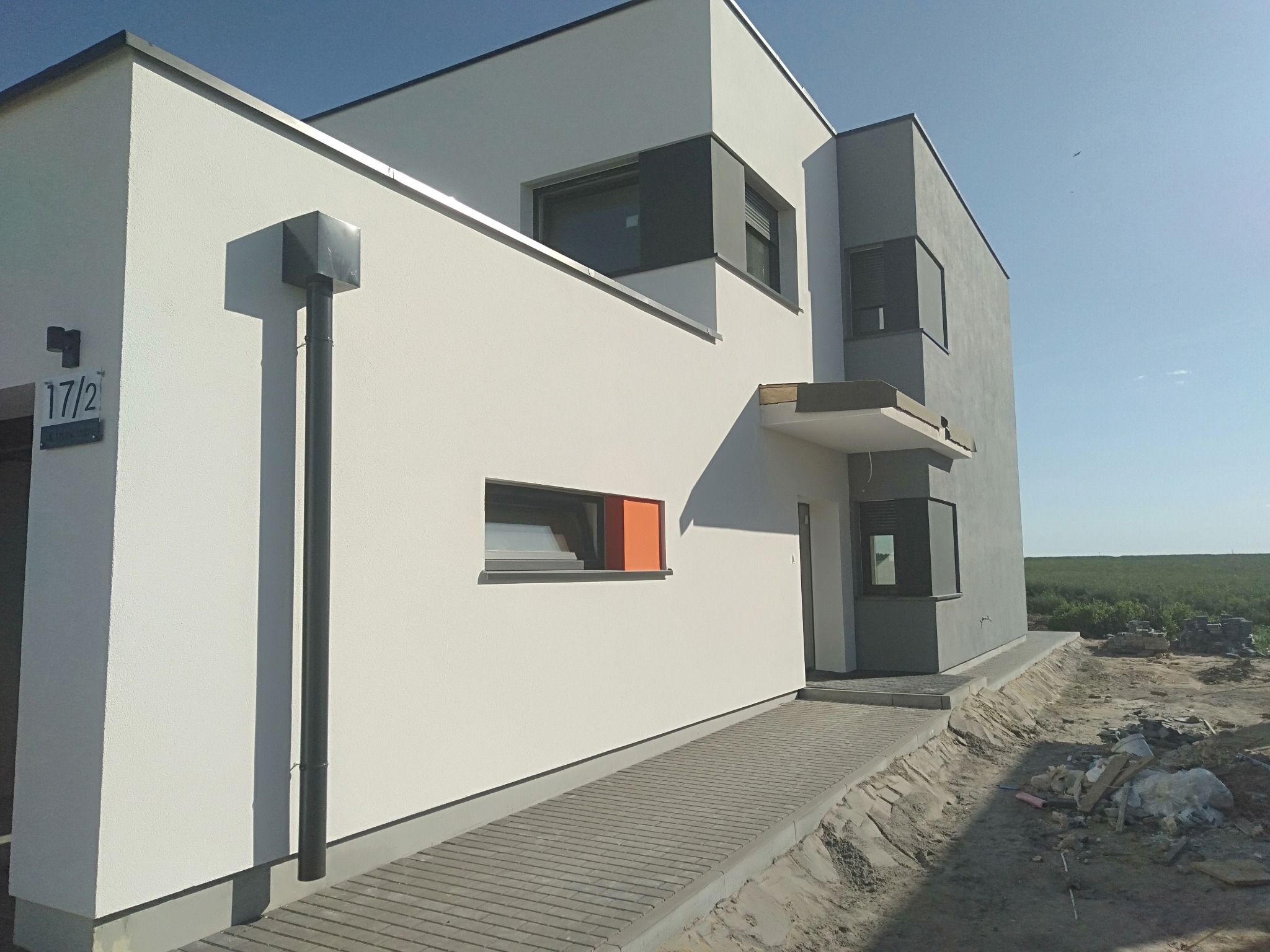 Nowoczesny dom jednorodzinny z białą elewacją i szarymi akcentami, widoczny z perspektywy chodnika z kostki brukowej, z numerem domu 17/2 na ścianie.