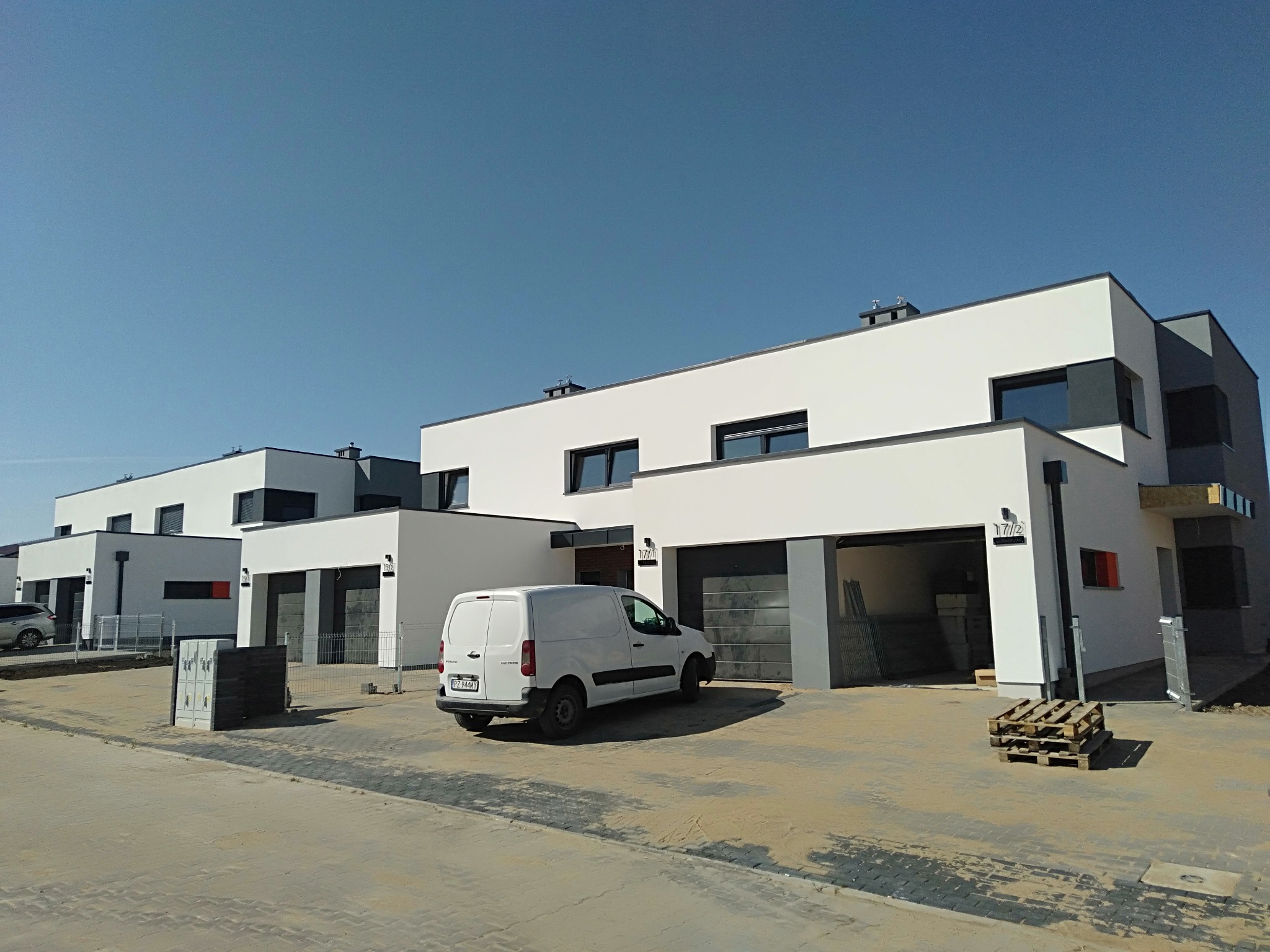 Nowoczesne szeregowe domy jednorodzinne z płaskimi dachami, białą elewacją i szarymi akcentami, częściowo z otwartymi garażami, biały samochód dostawczy na podjeździe, palety budowlane i kostka...