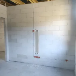 Surowy stan wnętrza z widocznymi przewodami elektrycznymi w ścianie z białego bloczka, puszki instalacyjne w kolorze pomarańczowym, sufit z drewnianą konstrukcją i zieloną membraną.