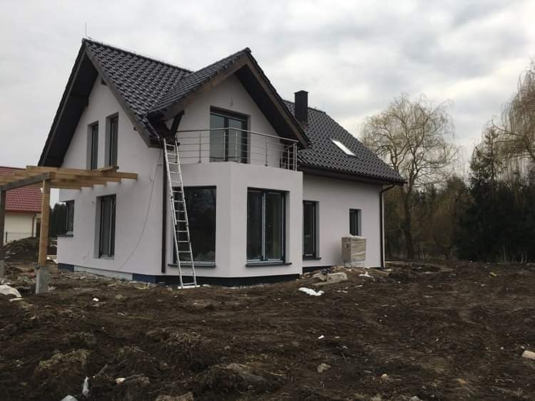 Nowoczesny dom jednorodzinny z białymi ścianami i ciemną dachówką, z balkonem z metalową balustradą, drabina oparta o ścianę, teren wokół domu w trakcie prac ziemnych.