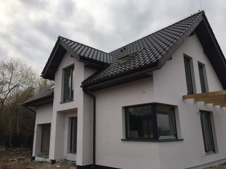 Nowoczesny dom jednorodzinny z elewacją w odcieniach szarości, czarną dachówką i grafitowymi oknami, widok z zewnątrz na tle pochmurnego nieba i drzew bez liści.