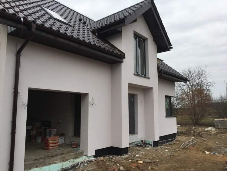 Nowoczesny dom jednorodzinny z jasną elewacją i ciemną dachówką, widok z zewnątrz na częściowo wykończony garaż z workami zaprawy i narzędziami, teren wokół domu w trakcie zagospodarowywania.