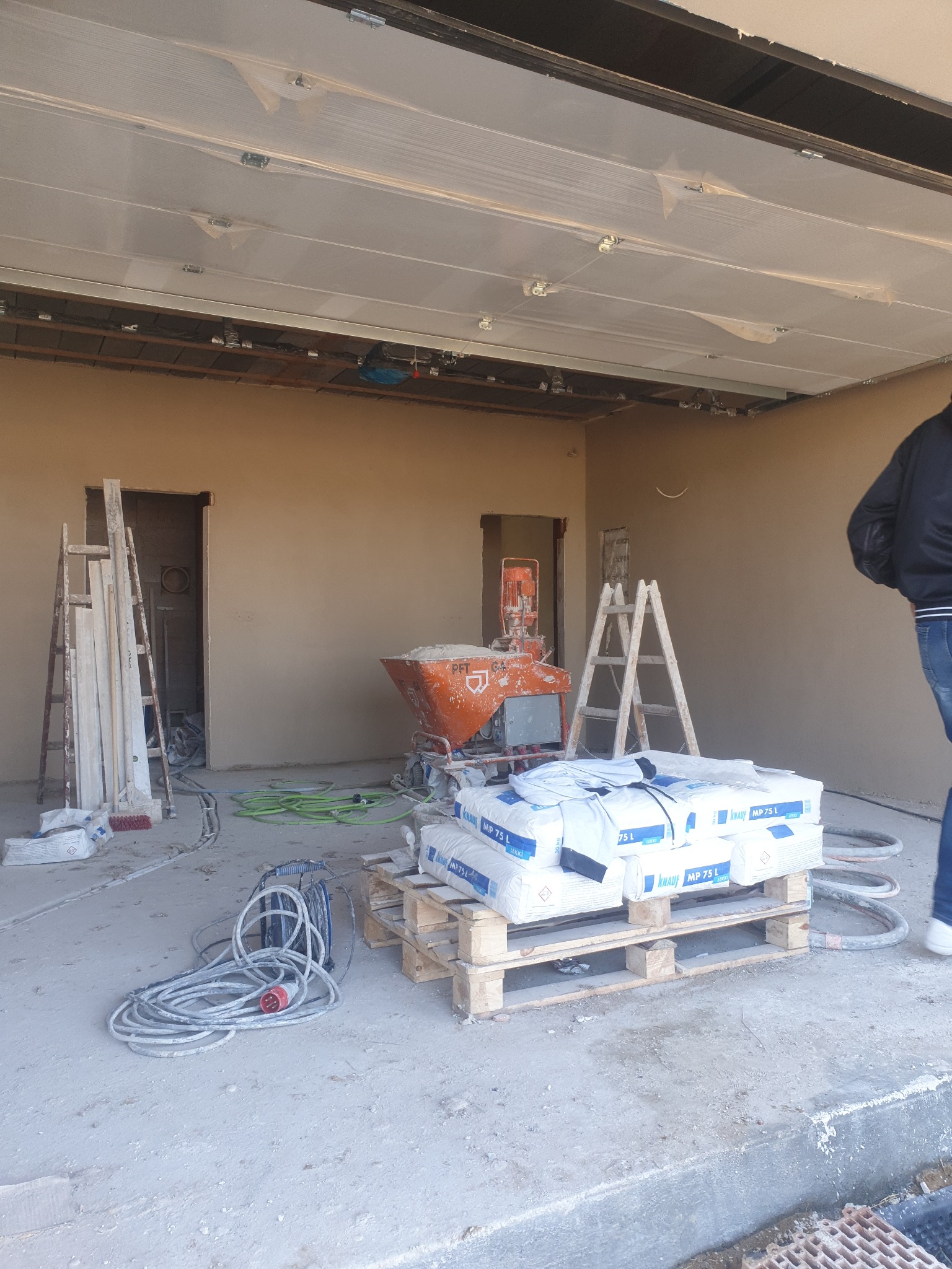 Wnętrze w trakcie prac tynkarskich: ściany pokryte świeżym tynkiem, maszyna tynkarska PFT, paleta z workami Knauf MP 75 L, drabiny i zwinięte węże na podłodze.