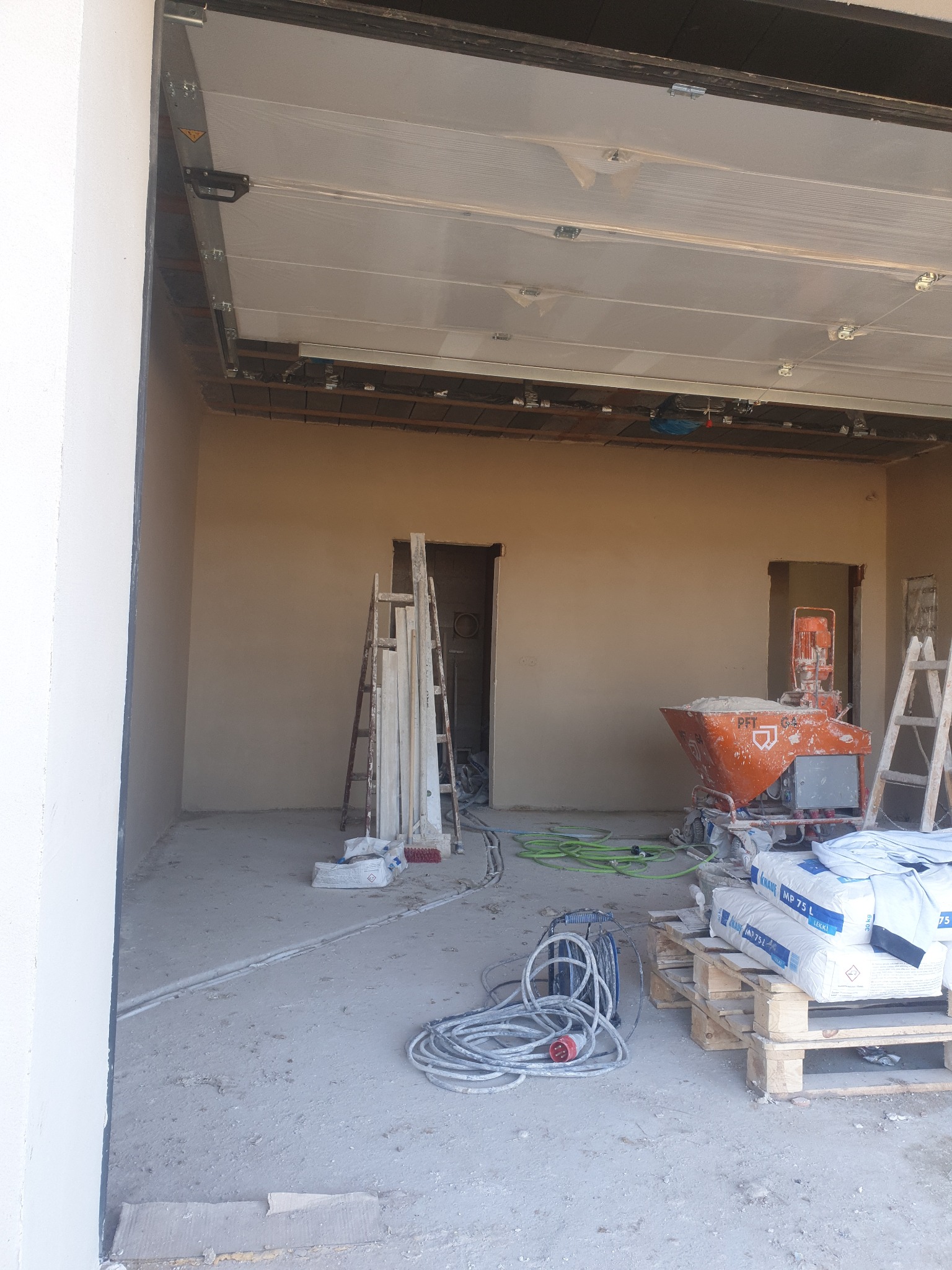 Wnętrze w trakcie prac wykończeniowych: ściany pokryte świeżym tynkiem, widoczna maszyna tynkarska PFT G4, worki z zaprawą Knauf MP 75 L na palecie, kable oraz drabina oparta o ścianę.