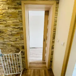 Wnętrze z drewnianą podłogą, ścianą wyłożoną kamieniem dekoracyjnym i otwartymi przesuwnymi drzwiami z drewnianą ościeżnicą, widoczna barierka zabezpieczająca schody.