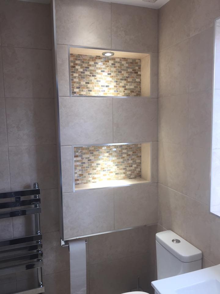 Wykończona łazienka z beżowymi płytkami, dekoracyjnymi wnękami z mozaiką i oświetleniem punktowym, widok na toaletę i grzejnik drabinkowy.
