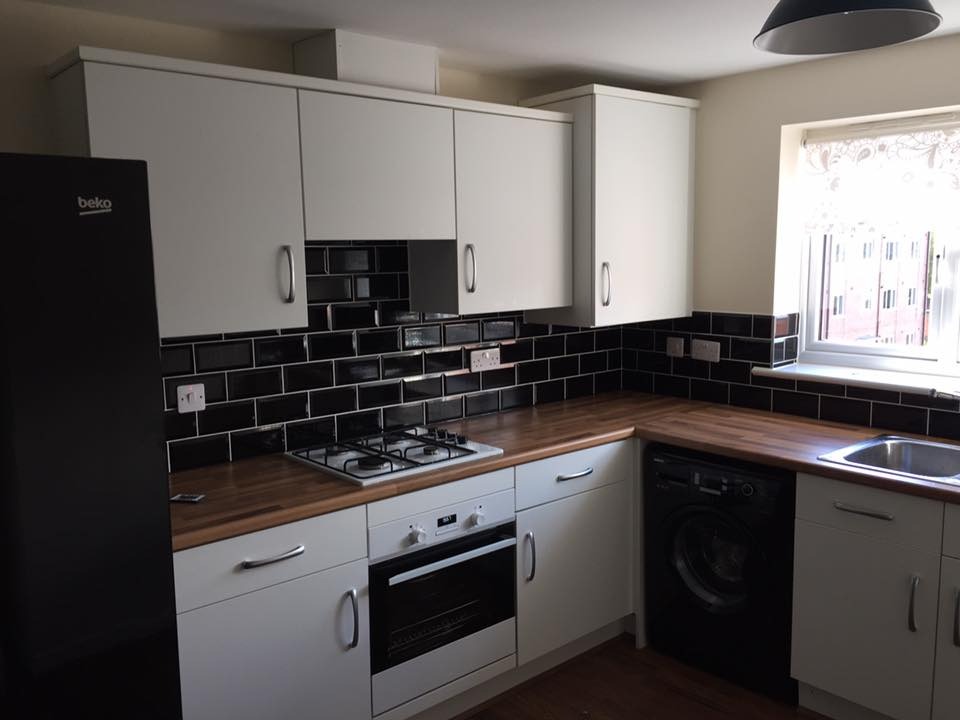 Wykończona kuchnia z białymi szafkami, drewnianym blatem i czarnymi płytkami na ścianie. Widoczny czarny piekarnik, czarna pralka i czarna lodówka Beko. Okno z firanką w tle.