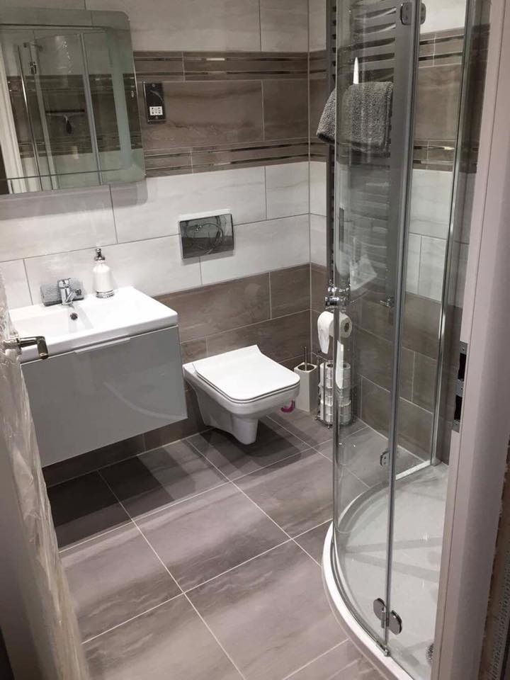 Nowoczesna łazienka z szaro-białymi płytkami imitującymi marmur, wisząca toaleta, umywalka z szafką, kabina prysznicowa z szarym ręcznikiem i srebrnymi akcesoriami.