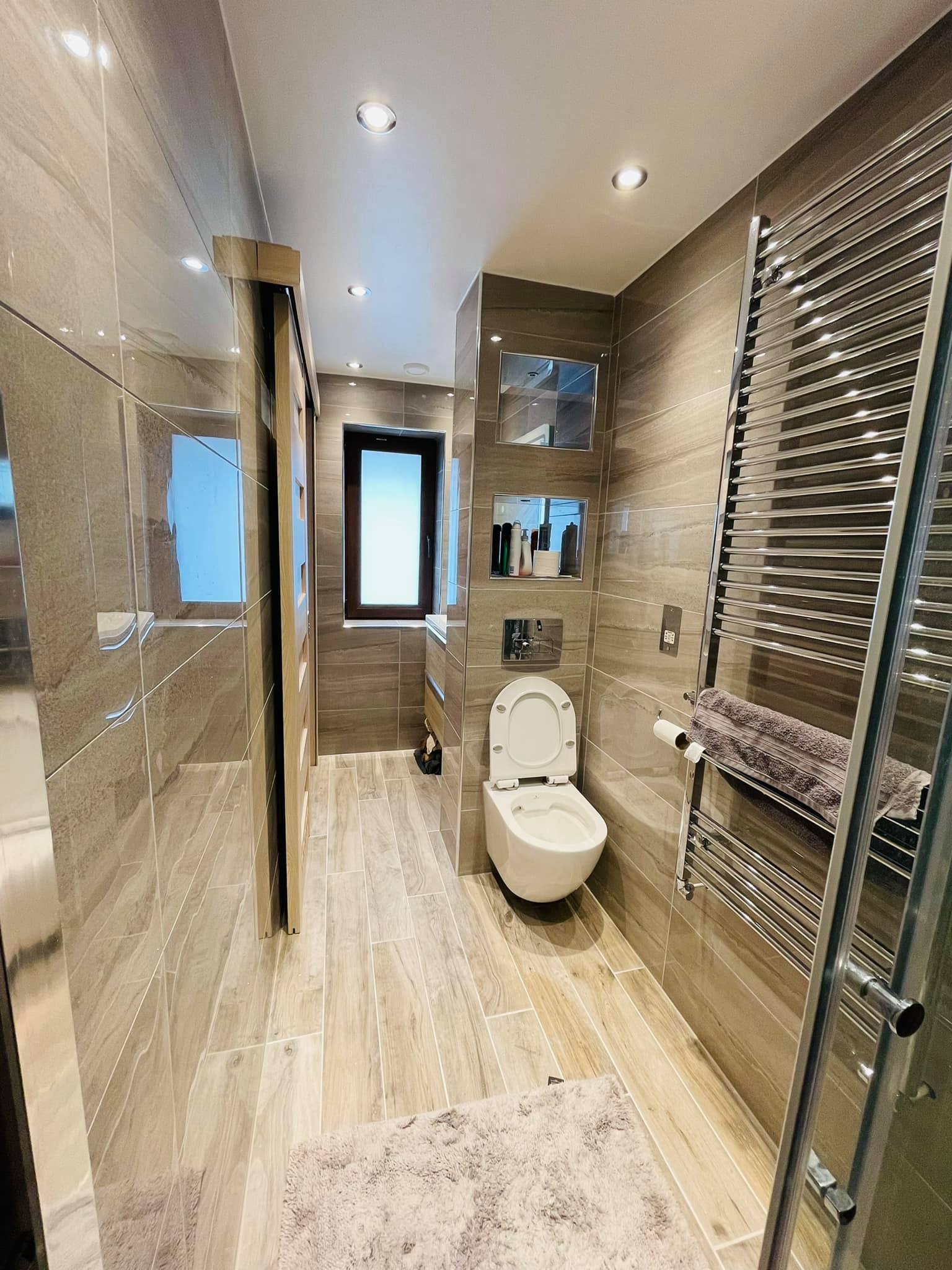 Wąska, nowoczesna łazienka z brązowymi płytkami imitującymi drewno, wiszącą toaletą z otwartą klapą, grzejnikiem drabinkowym z ręcznikiem i oknem w głębi.