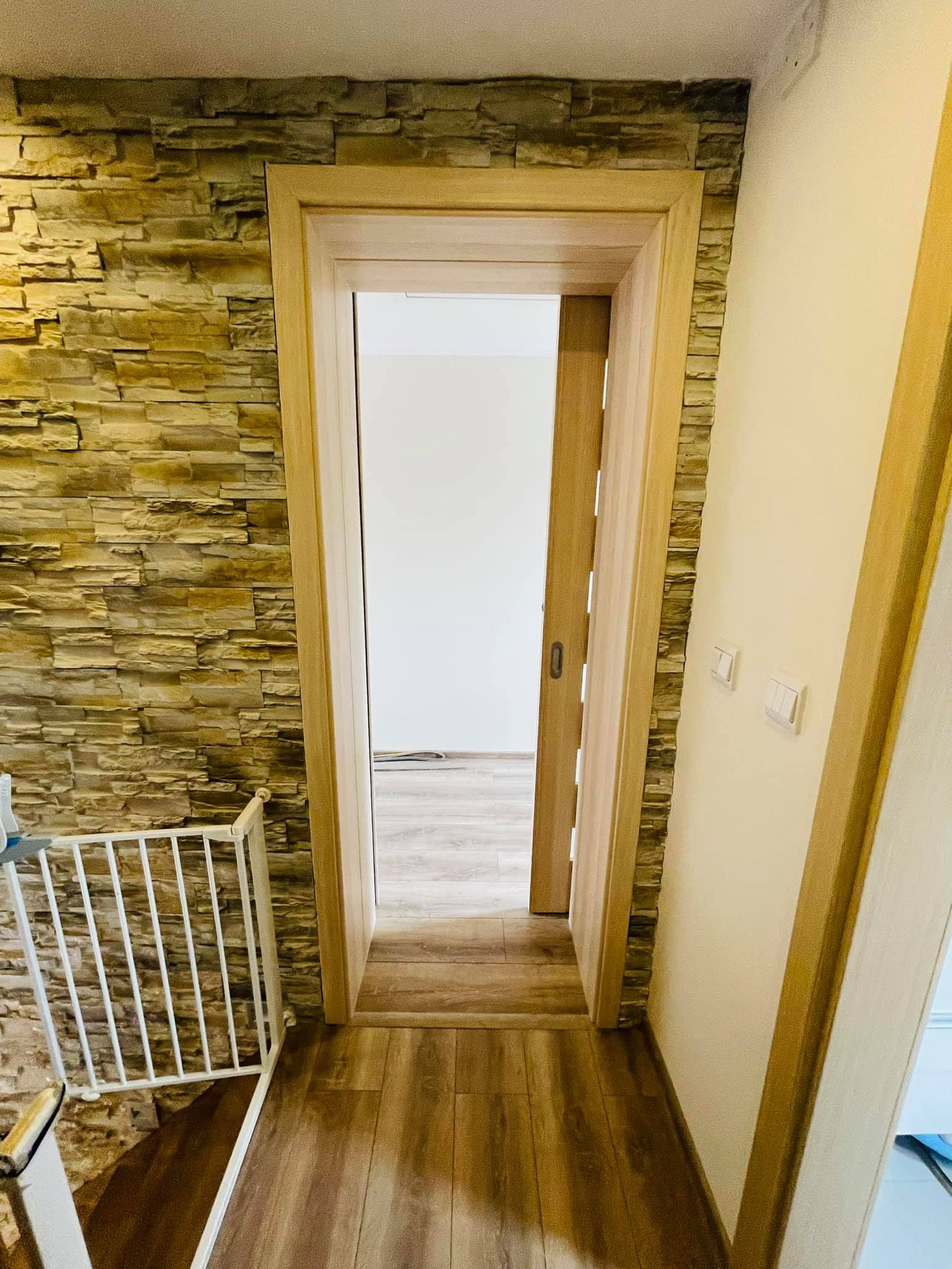 Wnętrze z drewnianą podłogą, ścianą wyłożoną kamieniem dekoracyjnym i otwartymi przesuwnymi drzwiami z drewnianą ościeżnicą, widoczna barierka zabezpieczająca schody.