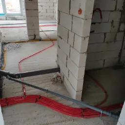 Surowy stan budowy: czerwone rury ogrzewania podłogowego, szare rury kanalizacyjne i ż&oacute;łte kable elektryczne prowadzone w betonie w nowym budynku z białych blok&oacute;w.