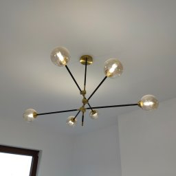 Montaż instalacji hydraulicznych i elektrycznych Mateusz Jaworski - Nowoczesny żyrandol sufitowy z sześcioma szklanymi kulami, czarne ramiona i złote elementy mocujące, widok z dołu na białym tle sufitu i ściany.