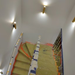 Montaż instalacji hydraulicznych i elektrycznych Mateusz Jaworski - Klatka schodowa w trakcie prac wykończeniowych, zabezpieczona folią malarską, widoczne nowoczesne, złote lampy ścienne.