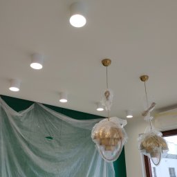 Montaż instalacji hydraulicznych i elektrycznych Mateusz Jaworski - Montaż oświetlenia sufitowego: złote lampy wiszące w folii ochronnej, białe reflektory punktowe, ściana zabezpieczona folią malarską, klimatyzator przy oknie.