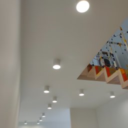 Montaż instalacji hydraulicznych i elektrycznych Mateusz Jaworski - Nowoczesne oświetlenie sufitowe z okrągłymi, białymi lampami LED w minimalistycznym wnętrzu, widok na schody z drewnianymi stopniami i zabezpieczeniami z taśmy, w tle widoczna pralka.