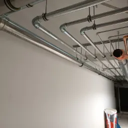 Różnorodne rury instalacyjne, w tym stalowe i miedziane, zamocowane pod sufitem w nowym budynku, widoczny worek z materiałem izolacyjnym Rockwool na podłodze, drabina w tle.