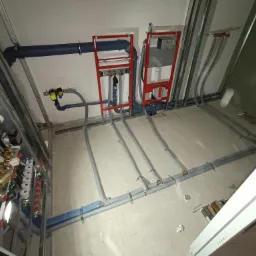 Surowe wnętrze łazienki w trakcie instalacji: widoczne rury, stelaże podtynkowe, metalowa konstrukcja mocująca, oraz rozdzielacz z zaworami, częściowo zasłonięty przez metalową ramę.