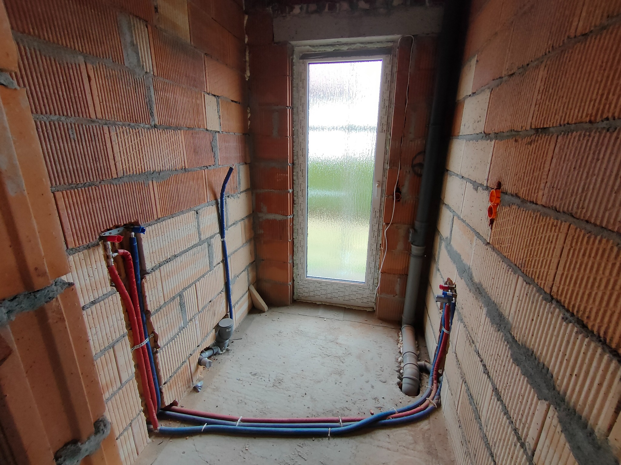 Surowy stan łazienki w trakcie prac hydraulicznych: widoczne rury doprowadzające wodę (czerwone i niebieskie) przy ścianie z cegły, pion kanalizacyjny i okno z szybą ornamentową.