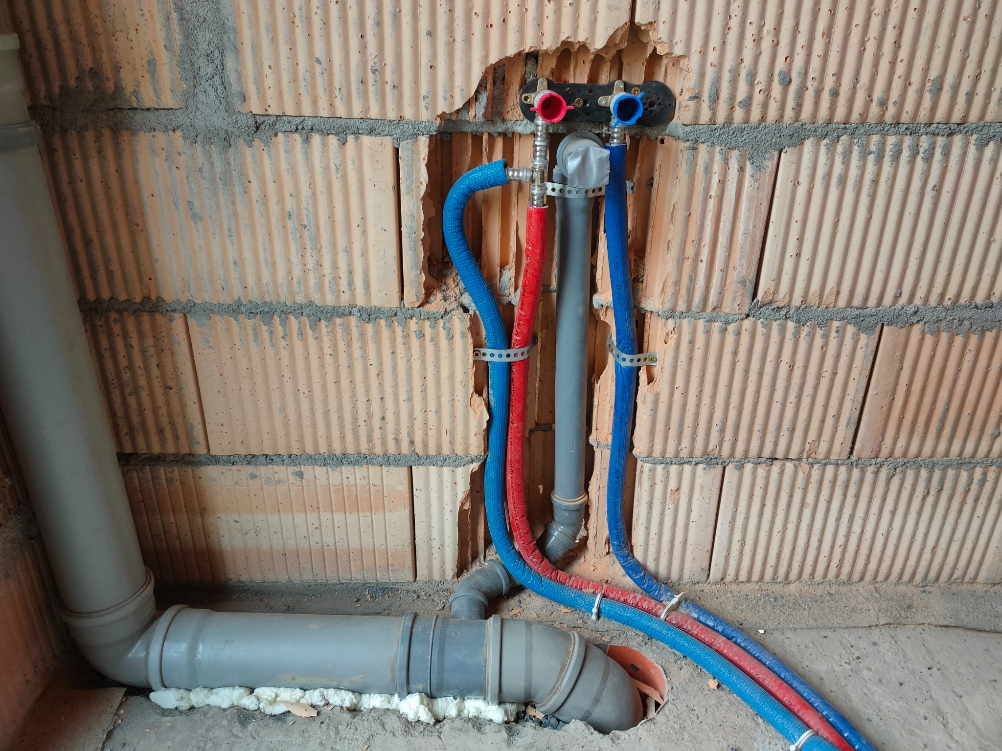 Wewnętrzna instalacja wodno-kanalizacyjna w trakcie montażu: rury PEX w kolorach czerwonym i niebieskim prowadzone wzdłuż surowej, ceglanej ściany, połączone z szarą rurą kanalizacyjną uszczelnioną...