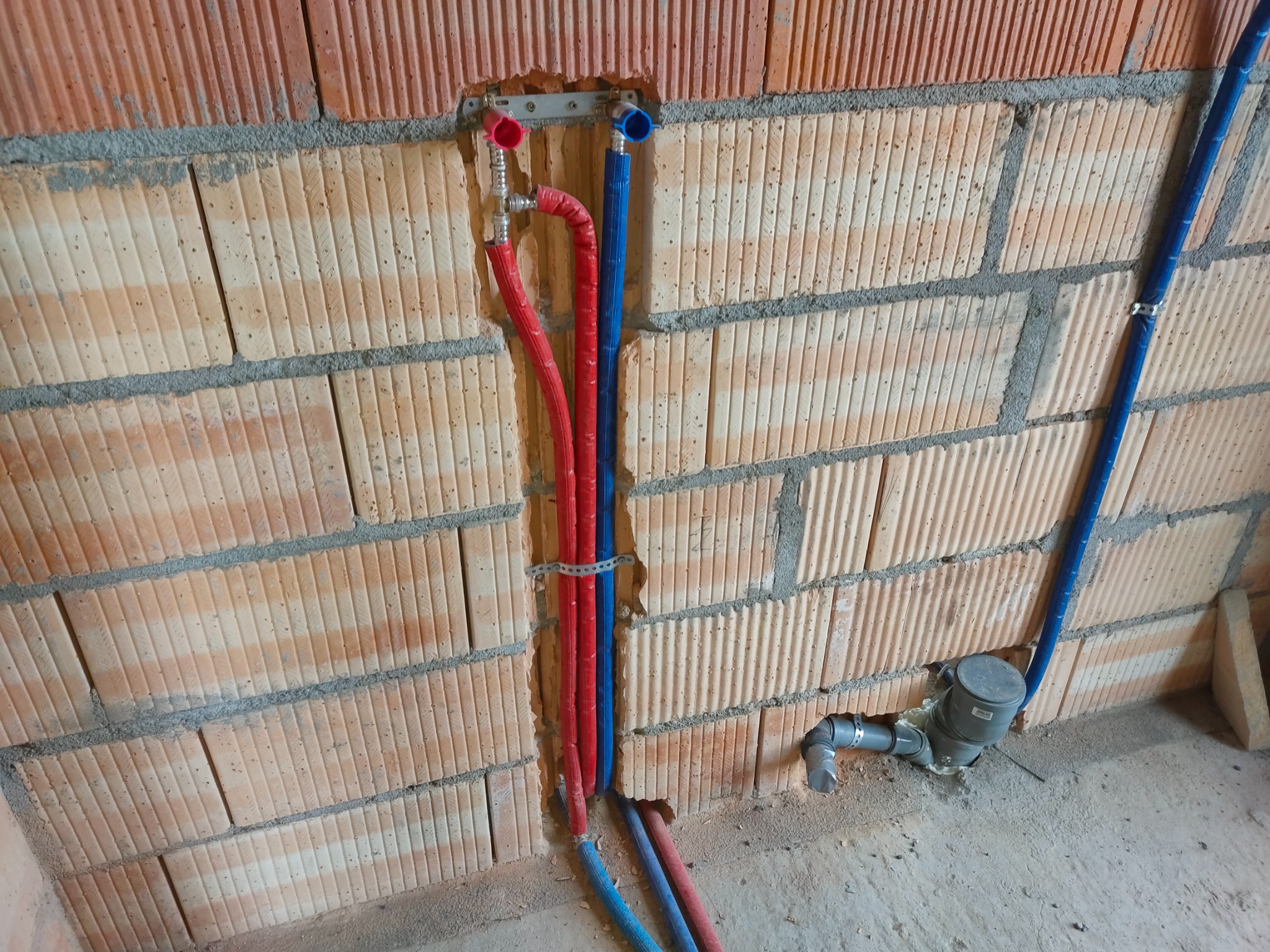 Czerwone i niebieskie rury wodne zamontowane w ścianie z cegły dziurawki, z widocznym licznikiem wody i fragmentem podłogi w surowym stanie.