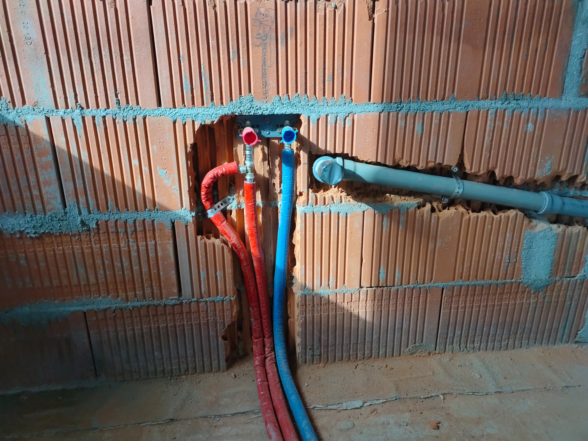 Czerwone i niebieskie rury instalacji wodnej wystające ze ściany z cegły dziurawki, widoczne fragmenty mocowań i szare rury odpływowe.