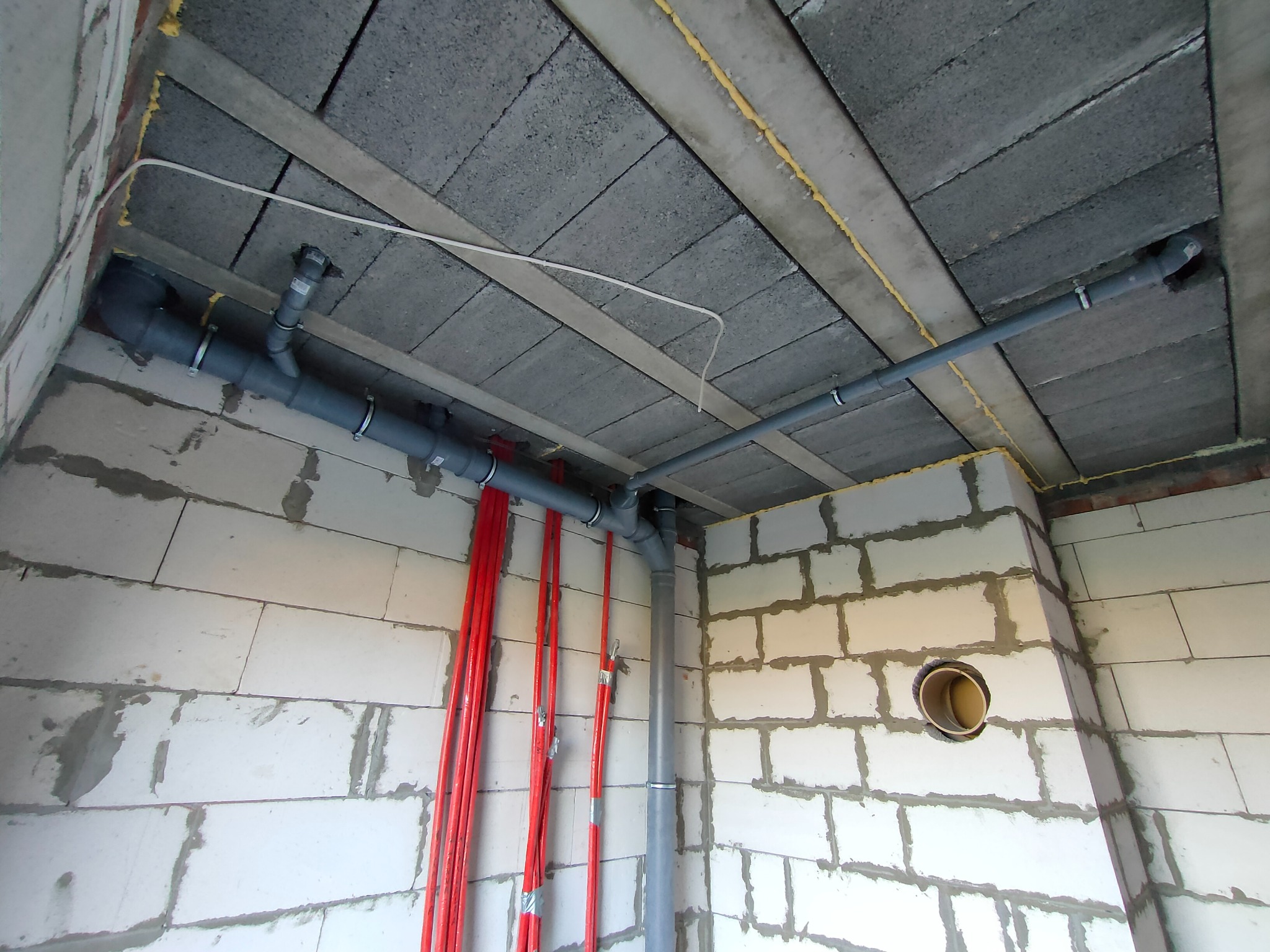 Szare rury kanalizacyjne pod stropem z betonowych bloczków, czerwone rury grzewcze przy ścianie z betonu komórkowego, widoczne mocowania i połączenia rur.