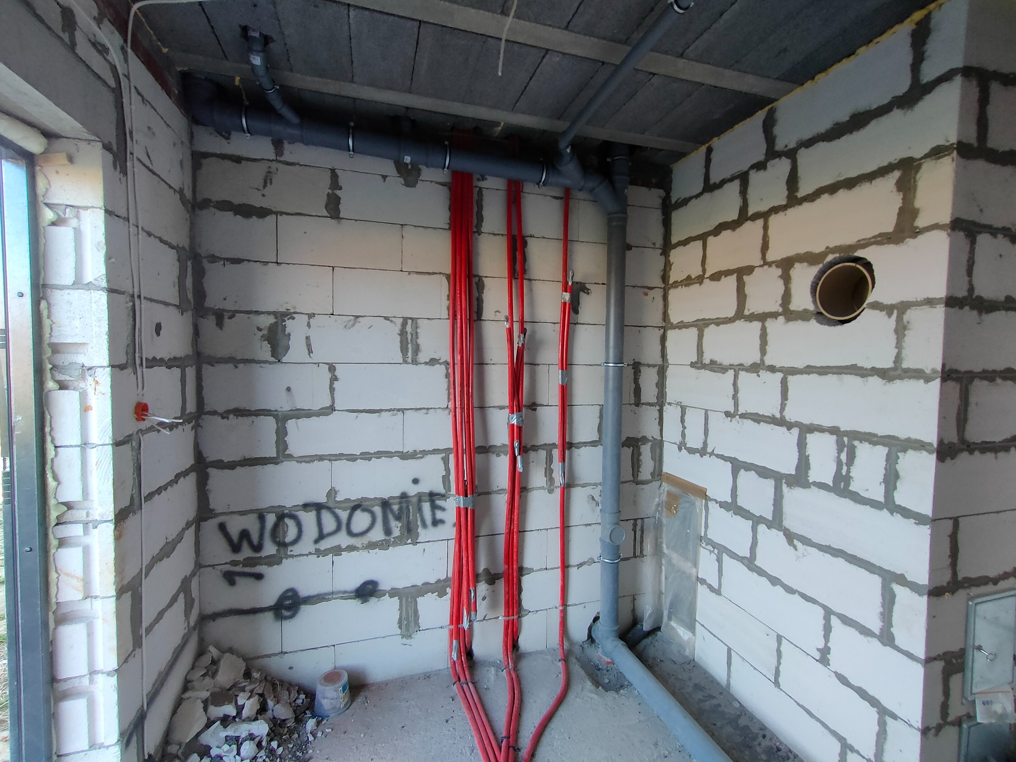 Surowy stan: czerwone rury instalacji ogrzewania podłogowego przymocowane taśmą do ściany z bloczków, szare rury kanalizacyjne pod stropem, widoczne napisy na ścianie i resztki materiałów budowlanych.