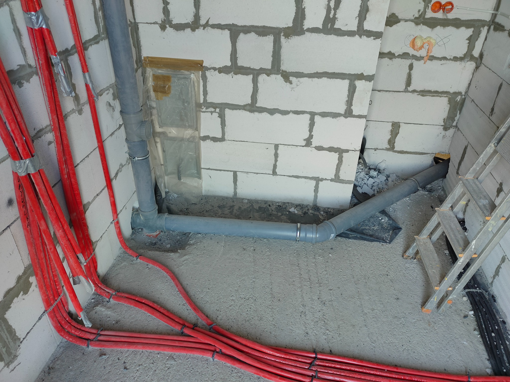 Wewnętrzne instalacje: Czerwone rury grzewcze prowadzone wzdłuż ściany z betonu komórkowego, szara rura kanalizacyjna, widoczne otwory na gniazdka elektryczne, drabina oparta o ścianę.