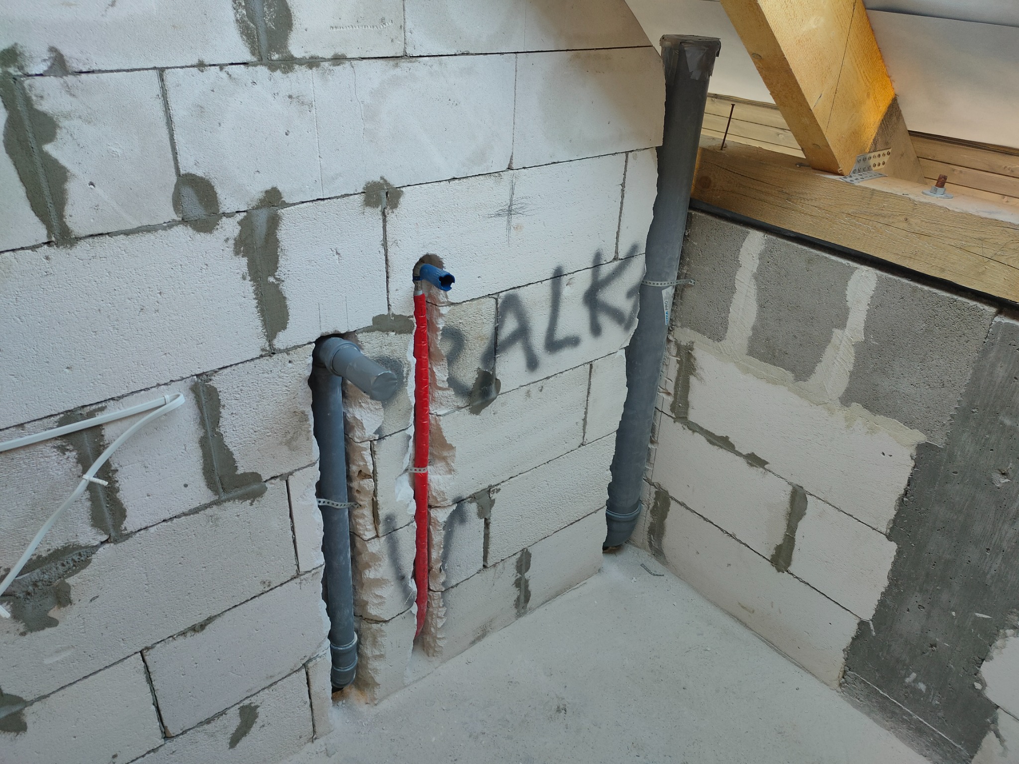 Surowa ściana z betonu komórkowego z pionowymi rurami instalacji wodnej i kanalizacyjnej, zabezpieczona czerwonym otuleniem, w trakcie montażu, z widocznym fragmentem drewnianej konstrukcji dachu.