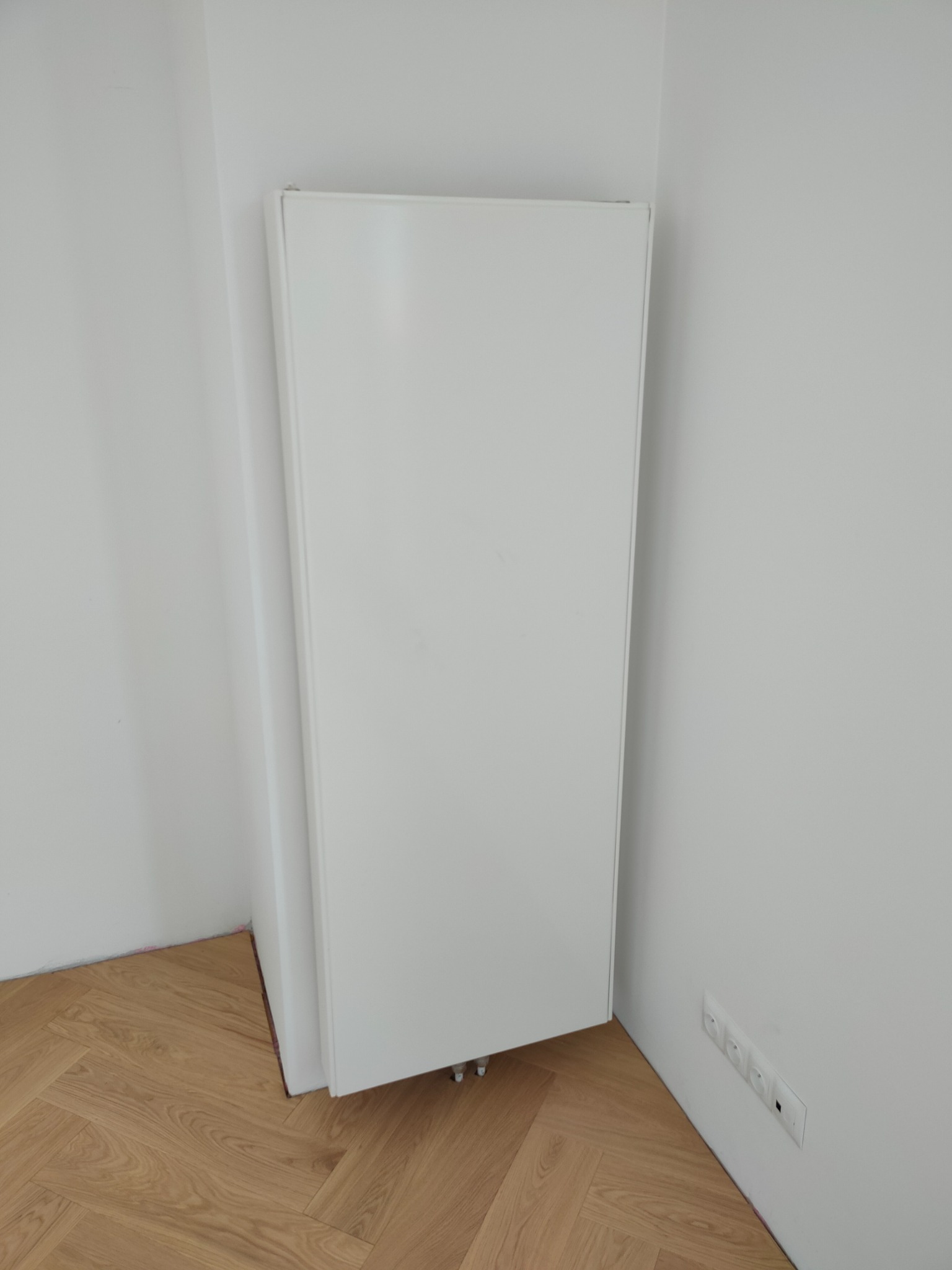 Biały, panelowy grzejnik zamontowany w rogu pomieszczenia z parkietem ułożonym w jodełkę, widoczne podłączenia do instalacji grzewczej i gniazdka elektryczne na ścianie.