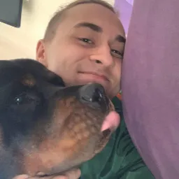 Mężczyzna z uśmiechem przytula rottweilera, który wystawia język, selfie w domowym otoczeniu.