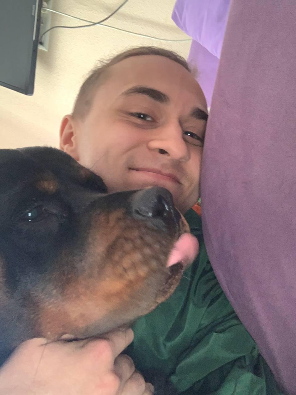 Mężczyzna z uśmiechem przytula rottweilera, który wystawia język, selfie w domowym otoczeniu.