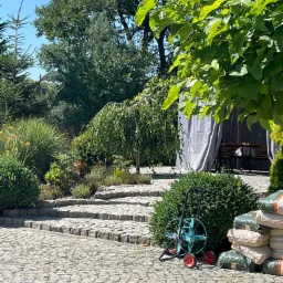 Ułożony z kostki brukowej podjazd do ogrodu z roślinami, drzewami, workami kory sosnowej i wózkiem na wąż ogrodowy