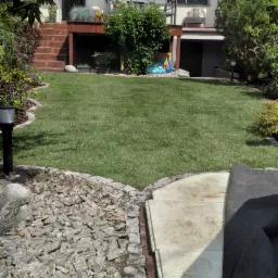 Świeżo założony trawnik z rolki przed domem, widoczne obrzeża z kamienia i kostki, w tle budynek mieszkalny z tarasem i balkonem.